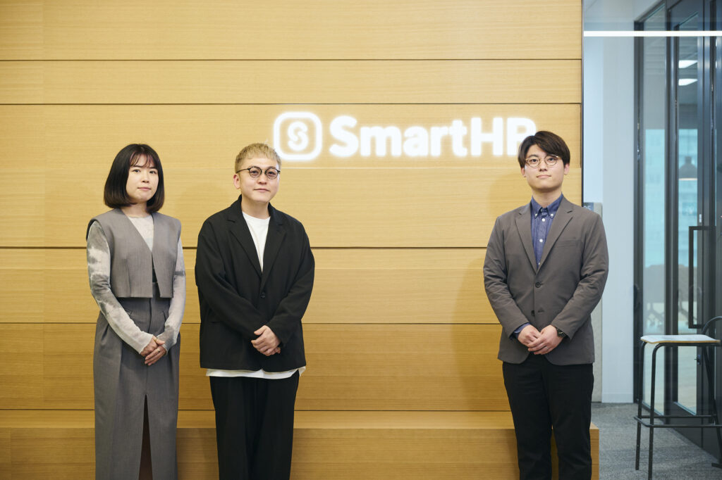 写真：SmartHRロゴをバックに並ぶ富士さん、山口さん、才流轟の3名