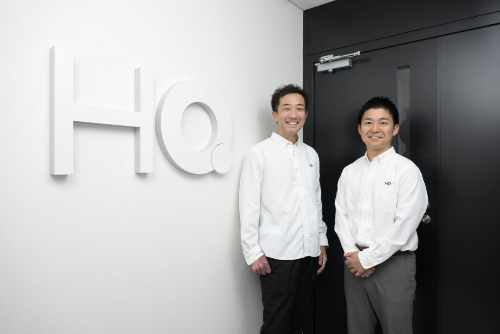 HQ稲垣さん、藤永さんがHQ社のロゴの前でほほ笑んでいる